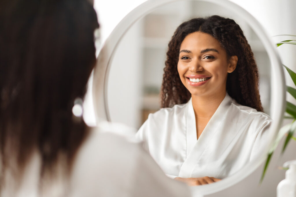 A young woman is looking in the mirror and smiling because she is more than satisfied with the results from her facial plastic surgery in Oklahoma City.