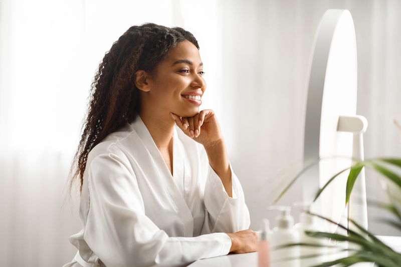 A young woman is looking in the mirror and smiling because she is more than satisfied with the results from her facial plastic surgery in Oklahoma City